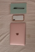 A light pink laptop with a metallic logo is placed on a fabric with a subtle pattern. Above the laptop, a smartphone with a white case and a pen on top of a light green notebook are arranged neatly.
