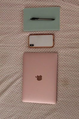 A light pink laptop with a metallic logo is placed on a fabric with a subtle pattern. Above the laptop, a smartphone with a white case and a pen on top of a light green notebook are arranged neatly.