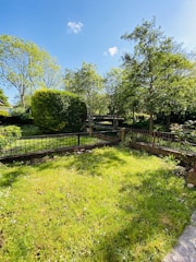 A lush green garden with a well-maintained lawn is bordered by a low brick and metal fence. The garden is adorned with a variety of trees and shrubs, providing ample shade. The sky is clear with a few fluffy white clouds, suggesting a bright and sunny day.