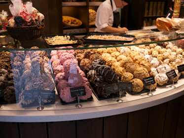 An assortment of pastries is displayed in a bakery showcase. The front row features various cookies and pastries, including chocolate and powdered sugar varieties. A wicker basket on the left contains wrapped baked goods. Behind the counter, a person in a white shirt and dark apron is working.