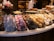 An assortment of pastries is displayed in a bakery showcase. The front row features various cookies and pastries, including chocolate and powdered sugar varieties. A wicker basket on the left contains wrapped baked goods. Behind the counter, a person in a white shirt and dark apron is working.