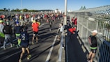 A large group of people participating in a marathon or running event on a bridge. Some runners are moving forward while others are stretching or preparing on the side of the path. The scene is brightly lit by sunlight, with a clear sky and an urban skyline visible in the background.