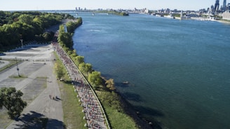 An aerial view of a large group of people participating in a marathon or race along a waterfront path. The path is lined with trees and runs parallel to a wide river with a cityscape visible in the distance. The scene is vibrant with green areas and blue water, and the participants are dressed in various colorful attire.