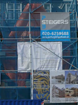 A construction site covered with blue scaffolding is visible. Several signs and banners advertising construction companies and services are attached to the scaffolding. There are details indicating restoration work being done, including contact information and project descriptions.