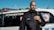 A man stands confidently in front of a police vehicle marked 'INTERVENTION', wearing a black uniform with a badge and a communication device. Several flags and solar panels can be seen in the background under a clear blue sky.
