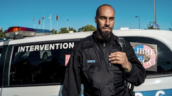 A man stands confidently in front of a police vehicle marked 'INTERVENTION', wearing a black uniform with a badge and a communication device. Several flags and solar panels can be seen in the background under a clear blue sky.