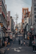 A bustling urban street scene with numerous shop signs and advertisements, written primarily in Korean, lining the sides of a narrow uphill street. There are a few pedestrians, including a person with a child, walking along the road. The buildings consist of various architectural styles and are closely packed together.
