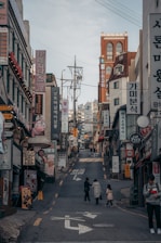 A bustling urban street scene with numerous shop signs and advertisements, written primarily in Korean, lining the sides of a narrow uphill street. There are a few pedestrians, including a person with a child, walking along the road. The buildings consist of various architectural styles and are closely packed together.