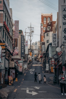 A bustling urban street scene with numerous shop signs and advertisements, written primarily in Korean, lining the sides of a narrow uphill street. There are a few pedestrians, including a person with a child, walking along the road. The buildings consist of various architectural styles and are closely packed together.