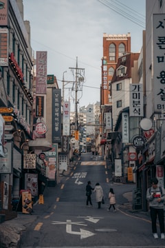 A bustling urban street scene with numerous shop signs and advertisements, written primarily in Korean, lining the sides of a narrow uphill street. There are a few pedestrians, including a person with a child, walking along the road. The buildings consist of various architectural styles and are closely packed together.