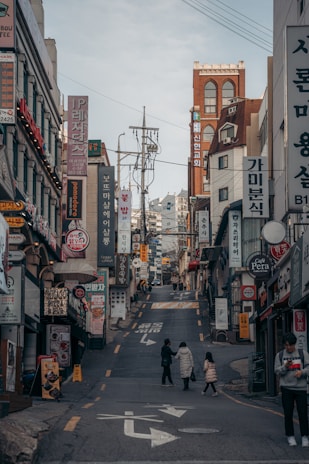 A bustling urban street scene with numerous shop signs and advertisements, written primarily in Korean, lining the sides of a narrow uphill street. There are a few pedestrians, including a person with a child, walking along the road. The buildings consist of various architectural styles and are closely packed together.