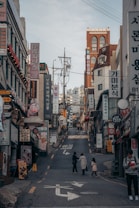 A bustling urban street scene with numerous shop signs and advertisements, written primarily in Korean, lining the sides of a narrow uphill street. There are a few pedestrians, including a person with a child, walking along the road. The buildings consist of various architectural styles and are closely packed together.