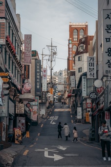 A bustling urban street scene with numerous shop signs and advertisements, written primarily in Korean, lining the sides of a narrow uphill street. There are a few pedestrians, including a person with a child, walking along the road. The buildings consist of various architectural styles and are closely packed together.