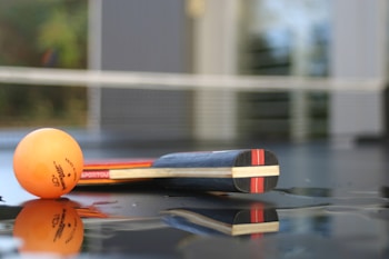 A table tennis racket and an orange ping pong ball are positioned on a reflective surface. The background includes a net and blurred elements, suggesting the setting is a ping pong table.