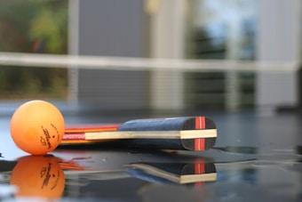 A table tennis racket and an orange ping pong ball are positioned on a reflective surface. The background includes a net and blurred elements, suggesting the setting is a ping pong table.