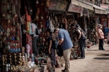 People are browsing a street market stall filled with an array of colorful handmade crafts, jewelry, and decorative items. The atmosphere is bustling, with several shoppers inspecting items while vendors display their goods. The market has a vibrant and traditional ambiance, with an assortment of brass items, woven textiles, and intricately designed artifacts.