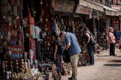 People are browsing a street market stall filled with an array of colorful handmade crafts, jewelry, and decorative items. The atmosphere is bustling, with several shoppers inspecting items while vendors display their goods. The market has a vibrant and traditional ambiance, with an assortment of brass items, woven textiles, and intricately designed artifacts.