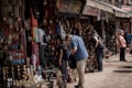 People are browsing a street market stall filled with an array of colorful handmade crafts, jewelry, and decorative items. The atmosphere is bustling, with several shoppers inspecting items while vendors display their goods. The market has a vibrant and traditional ambiance, with an assortment of brass items, woven textiles, and intricately designed artifacts.