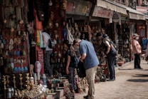 People are browsing a street market stall filled with an array of colorful handmade crafts, jewelry, and decorative items. The atmosphere is bustling, with several shoppers inspecting items while vendors display their goods. The market has a vibrant and traditional ambiance, with an assortment of brass items, woven textiles, and intricately designed artifacts.