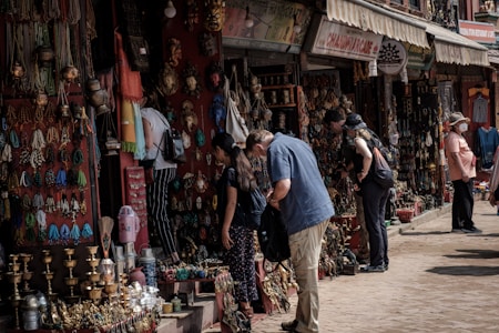 People are browsing a street market stall filled with an array of colorful handmade crafts, jewelry, and decorative items. The atmosphere is bustling, with several shoppers inspecting items while vendors display their goods. The market has a vibrant and traditional ambiance, with an assortment of brass items, woven textiles, and intricately designed artifacts.