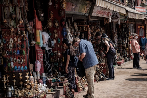 People are browsing a street market stall filled with an array of colorful handmade crafts, jewelry, and decorative items. The atmosphere is bustling, with several shoppers inspecting items while vendors display their goods. The market has a vibrant and traditional ambiance, with an assortment of brass items, woven textiles, and intricately designed artifacts.