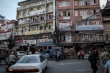 A bustling urban street scene with multi-story buildings featuring colorful signage advertising various businesses, including a spa, tattoo inn, and handicraft store. Numerous tangled electrical wires run across the buildings. People are walking along the sidewalk, and vehicles, including a car and a motorbike, are present on the street.