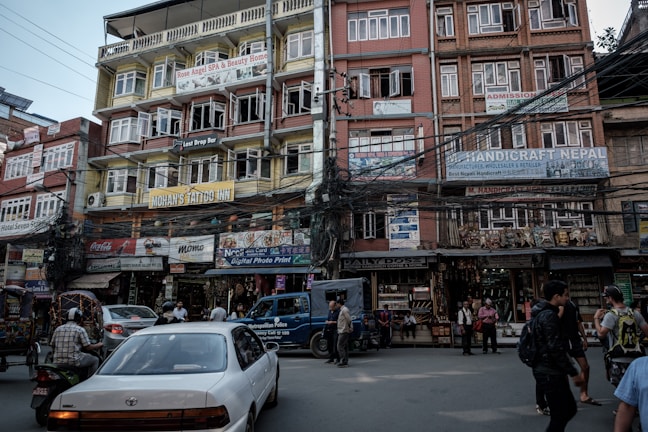 A bustling urban street scene with multi-story buildings featuring colorful signage advertising various businesses, including a spa, tattoo inn, and handicraft store. Numerous tangled electrical wires run across the buildings. People are walking along the sidewalk, and vehicles, including a car and a motorbike, are present on the street.