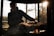 A silhouette of a person is seen behind a grill, with several pizzas cooking on it. The scene is outdoors, with trees visible in the background. Sunlight filters through the trees, casting a warm glow over the scene.