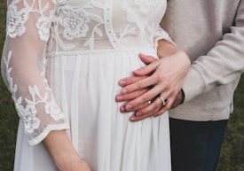 A close-up of a couple with hands placed affectionately on a pregnant belly. The woman is wearing a white, lace dress with floral patterns. The man's arm is draped around her from behind, and both wear wedding rings.