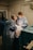 Two medical professionals are performing surgery in an operating room. They are dressed in blue surgical gowns, masks, and hairnets. Surgical instruments and equipment are placed on a table nearby, and the scene is illuminated by focused lighting.