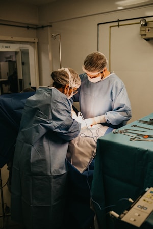 Two medical professionals are performing surgery in an operating room. They are dressed in blue surgical gowns, masks, and hairnets. Surgical instruments and equipment are placed on a table nearby, and the scene is illuminated by focused lighting.