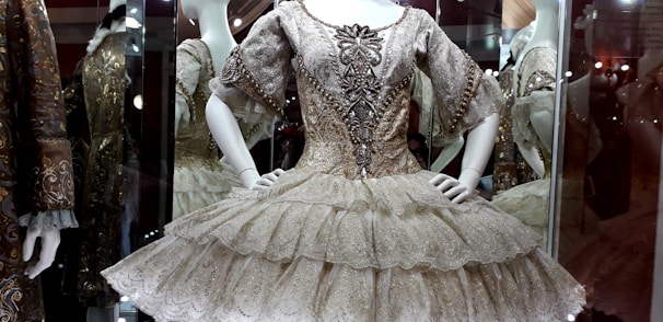A detailed and ornate ballet costume is displayed in a glass case. The dress features intricate embroidery with metallic embellishments on the bodice and sleeves, and a layered, ruffled skirt. Mannequins in the background are also dressed in similarly elaborate outfits, suggesting an exhibition or museum setting.