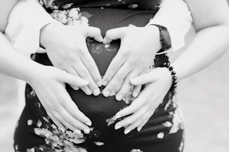 Two pairs of hands gently rest on a pregnant belly, forming a heart shape with the fingers. The person is wearing a floral dress, and a watch and bracelet can be seen on the wrists of one pair of hands.