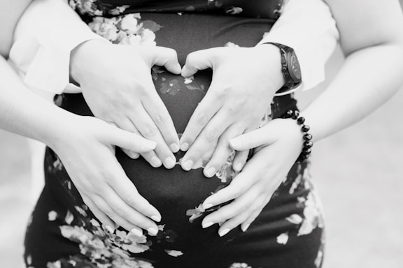 Two pairs of hands gently rest on a pregnant belly, forming a heart shape with the fingers. The person is wearing a floral dress, and a watch and bracelet can be seen on the wrists of one pair of hands.