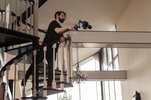 A person wearing dark clothing and a face mask is leaning over a spiraling staircase railing, holding a professional camera setup. The interior setting features a high ceiling with large windows, allowing natural light to illuminate the space. Elegant chandeliers hang from the ceiling, and the decor suggests a modern, sophisticated style.