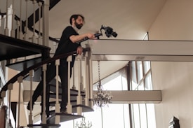 A person wearing dark clothing and a face mask is leaning over a spiraling staircase railing, holding a professional camera setup. The interior setting features a high ceiling with large windows, allowing natural light to illuminate the space. Elegant chandeliers hang from the ceiling, and the decor suggests a modern, sophisticated style.