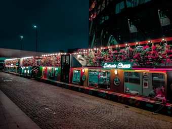 A vibrant train carriage is decorated with colorful flowers and lights on the exterior, creating a festive and inviting atmosphere. The interior is visible through large windows, showing people seated inside. The setting is on a cobblestone platform at night, with low light adding to the charming and cozy ambiance.