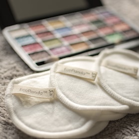 Three cream-colored reusable cotton pads with the label 'EcoPanda™' are placed on a soft, textured surface. In the background, a palette of watercolors or makeup shows assorted hues, slightly out of focus.