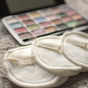 Three cream-colored reusable cotton pads with the label 'EcoPanda™' are placed on a soft, textured surface. In the background, a palette of watercolors or makeup shows assorted hues, slightly out of focus.