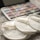 Three cream-colored reusable cotton pads with the label 'EcoPanda™' are placed on a soft, textured surface. In the background, a palette of watercolors or makeup shows assorted hues, slightly out of focus.