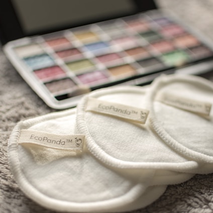 Three cream-colored reusable cotton pads with the label 'EcoPanda™' are placed on a soft, textured surface. In the background, a palette of watercolors or makeup shows assorted hues, slightly out of focus.