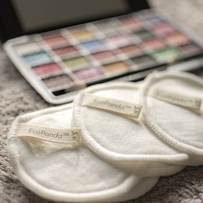 Three cream-colored reusable cotton pads with the label 'EcoPanda™' are placed on a soft, textured surface. In the background, a palette of watercolors or makeup shows assorted hues, slightly out of focus.