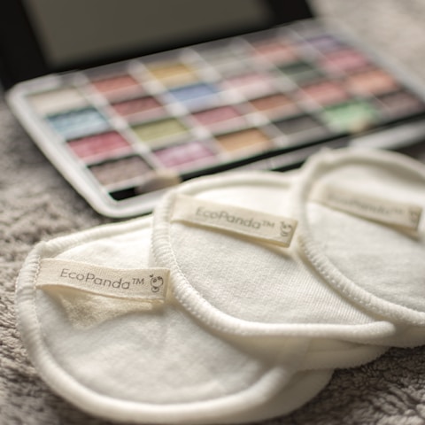 Three cream-colored reusable cotton pads with the label 'EcoPanda™' are placed on a soft, textured surface. In the background, a palette of watercolors or makeup shows assorted hues, slightly out of focus.
