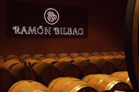 A dimly lit room filled with rows of wooden wine barrels, each marked with the brand name. A large illuminated sign displaying 'Ramón Bilbao' is mounted on the wall above the barrels, adding to the atmosphere.