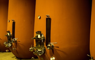 Large, cylindrical vessels with metal fixtures are aligned side by side. The tanks have a matte orange-brown finish, with stainless steel taps and dials attached near the bottom. The setting appears industrial or related to beverage production or storage.