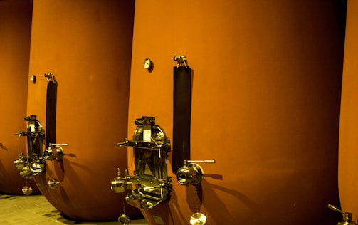 Large, cylindrical vessels with metal fixtures are aligned side by side. The tanks have a matte orange-brown finish, with stainless steel taps and dials attached near the bottom. The setting appears industrial or related to beverage production or storage.