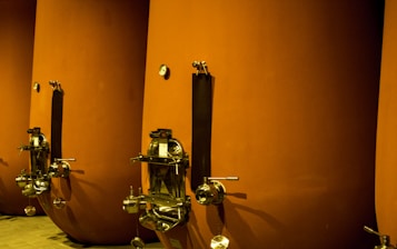 Large, cylindrical vessels with metal fixtures are aligned side by side. The tanks have a matte orange-brown finish, with stainless steel taps and dials attached near the bottom. The setting appears industrial or related to beverage production or storage.