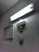 A rectangular fluorescent light fixture is mounted on a textured wall, casting bright illumination. Below the light, a rectangular speaker or buzzer and a wall switch are visible. To the side, a ventilation grille is attached to the wall.