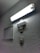 A rectangular fluorescent light fixture is mounted on a textured wall, casting bright illumination. Below the light, a rectangular speaker or buzzer and a wall switch are visible. To the side, a ventilation grille is attached to the wall.