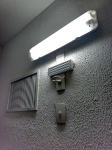 A rectangular fluorescent light fixture is mounted on a textured wall, casting bright illumination. Below the light, a rectangular speaker or buzzer and a wall switch are visible. To the side, a ventilation grille is attached to the wall.